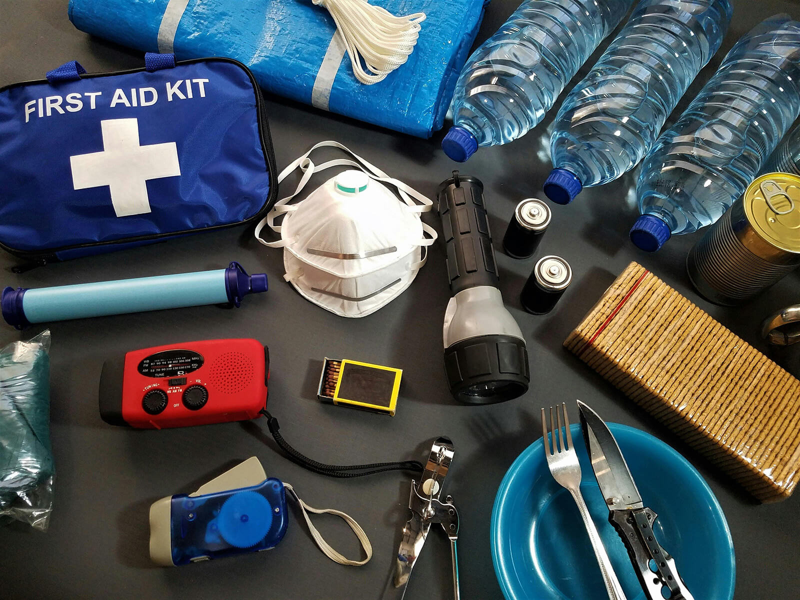 emergency items on table: Mask, utenselce, mask, matches, canned food, crackers, water, flash lights, radio, trap, first aid kit